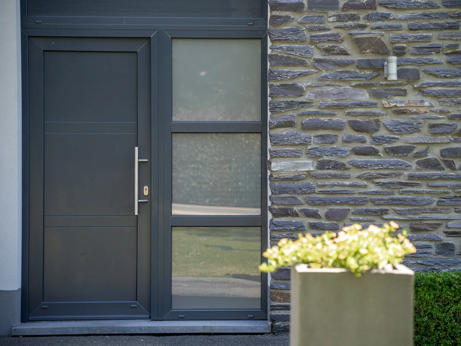 Porte d'entrée anthracite à imposte vitrée, encastrée dans un parement de schiste appareillé à sec